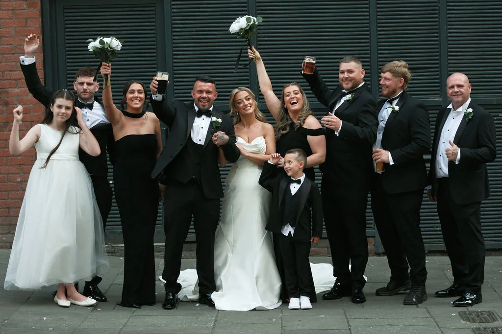 A joyful wedding party in black tie attire poses together on a paved street in front of a dark slatted wall, raising drinks and bouquets in celebration.