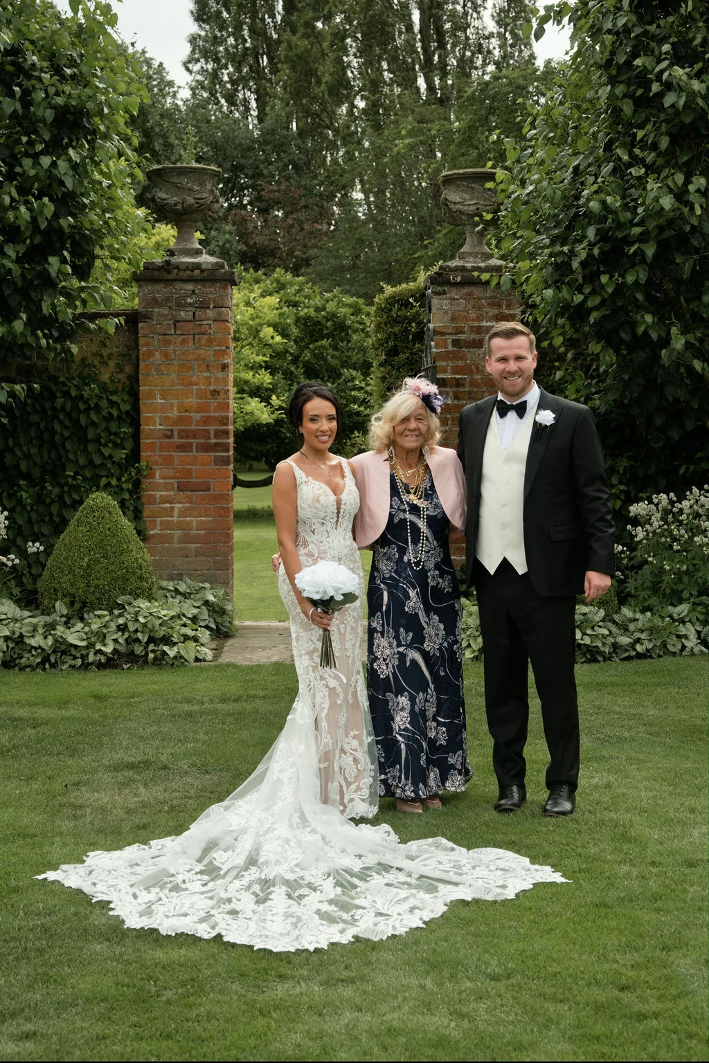 A bride in a lace mermaid gown and a groom in a classic tuxedo pose with an elderly family member in a manicured garden setting.