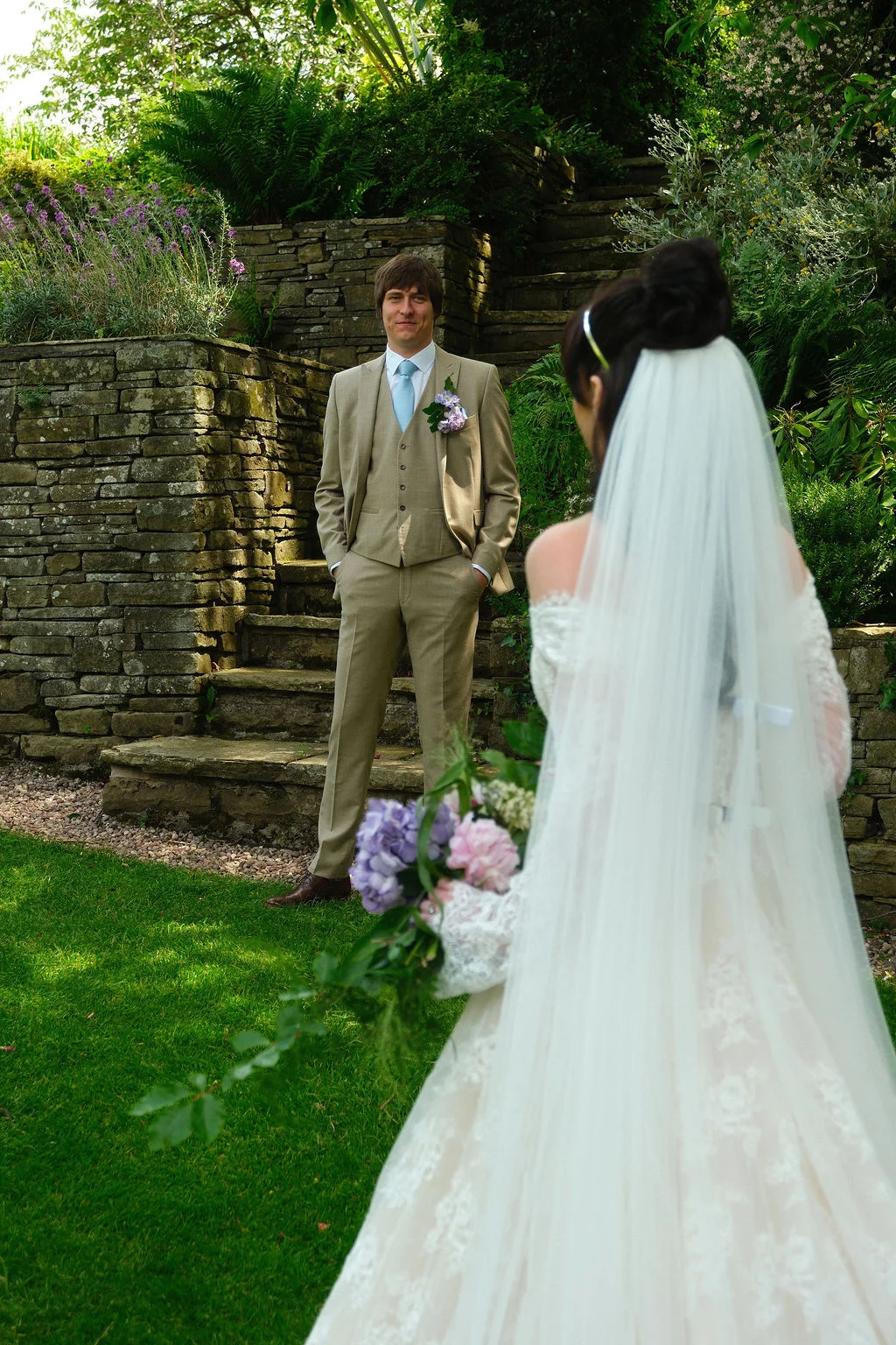 A groom in a beige three piece suit smiles as he sees his bride wearing a lace gown and long veil in a garden.