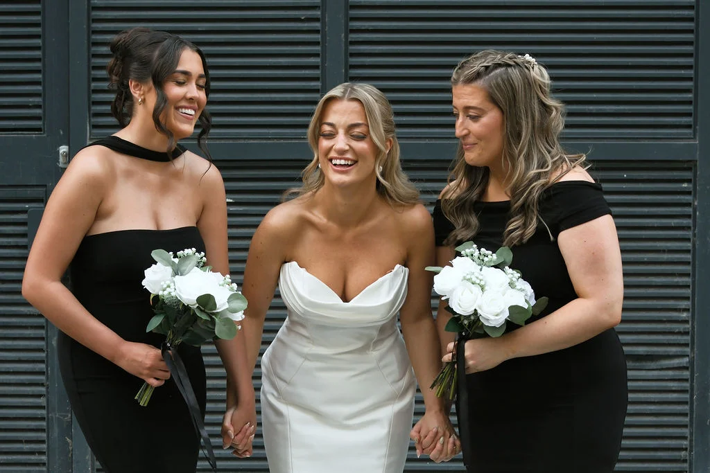 A joyful bride in a white strapless satin gown laughs heartily while holding hands with two bridesmaids wearing elegant black dresses and holding white rose bouquets.