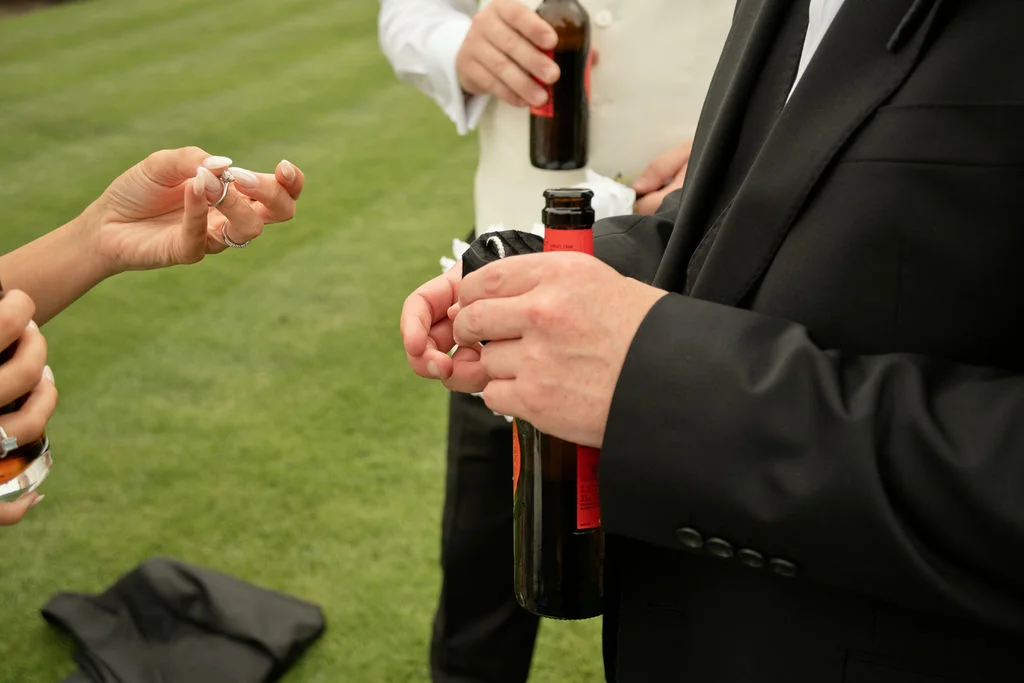 A guest shows off her engagement ring while chatting with friends holding bottles of beer during a wedding drinks reception outdoors.