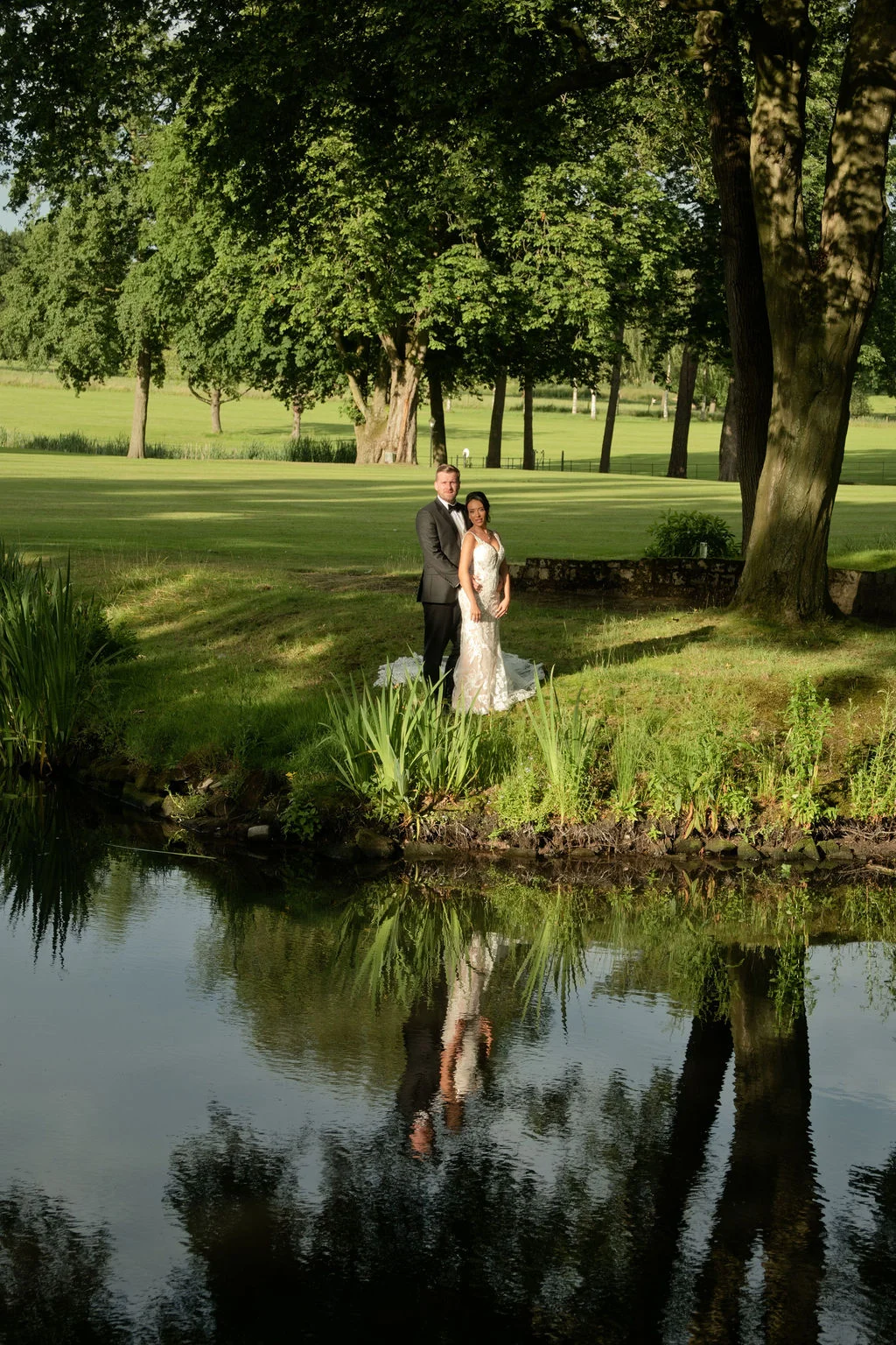 Newlyweds pose by a tranquil lake in an expansive manicured garden, creating a stunning reflection in the water under bright sunlight.