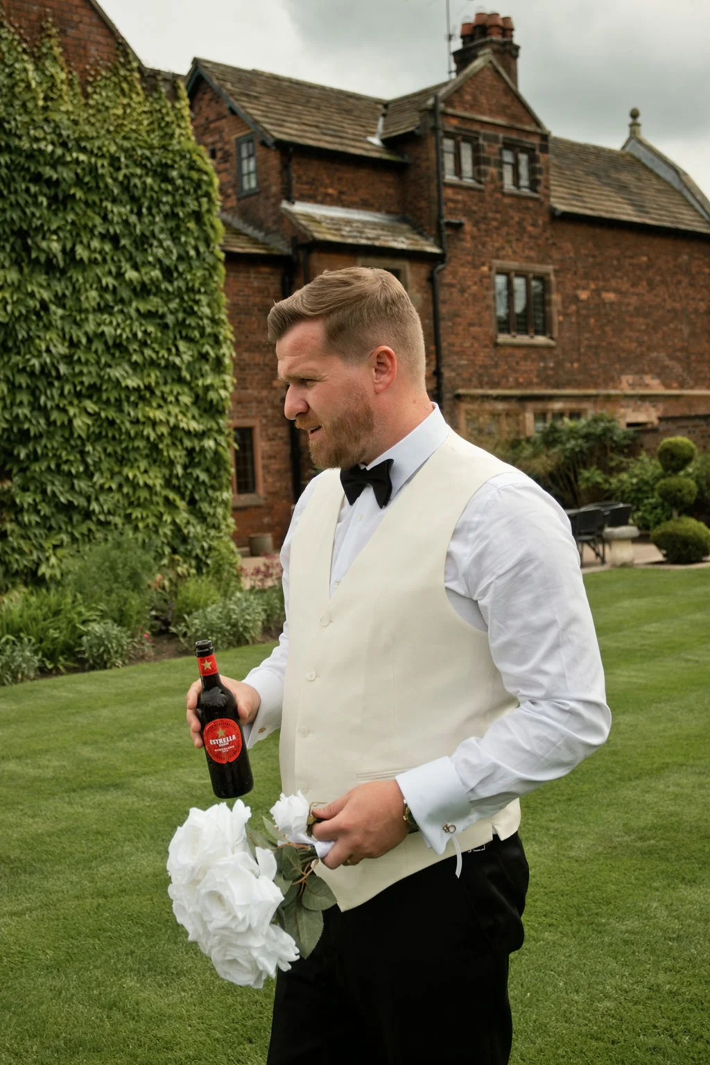 A groom stands on a manicured lawn outside a historic brick country house, wearing a cream waistcoat, white shirt, and black bow tie while holding a beer bottle and white roses.