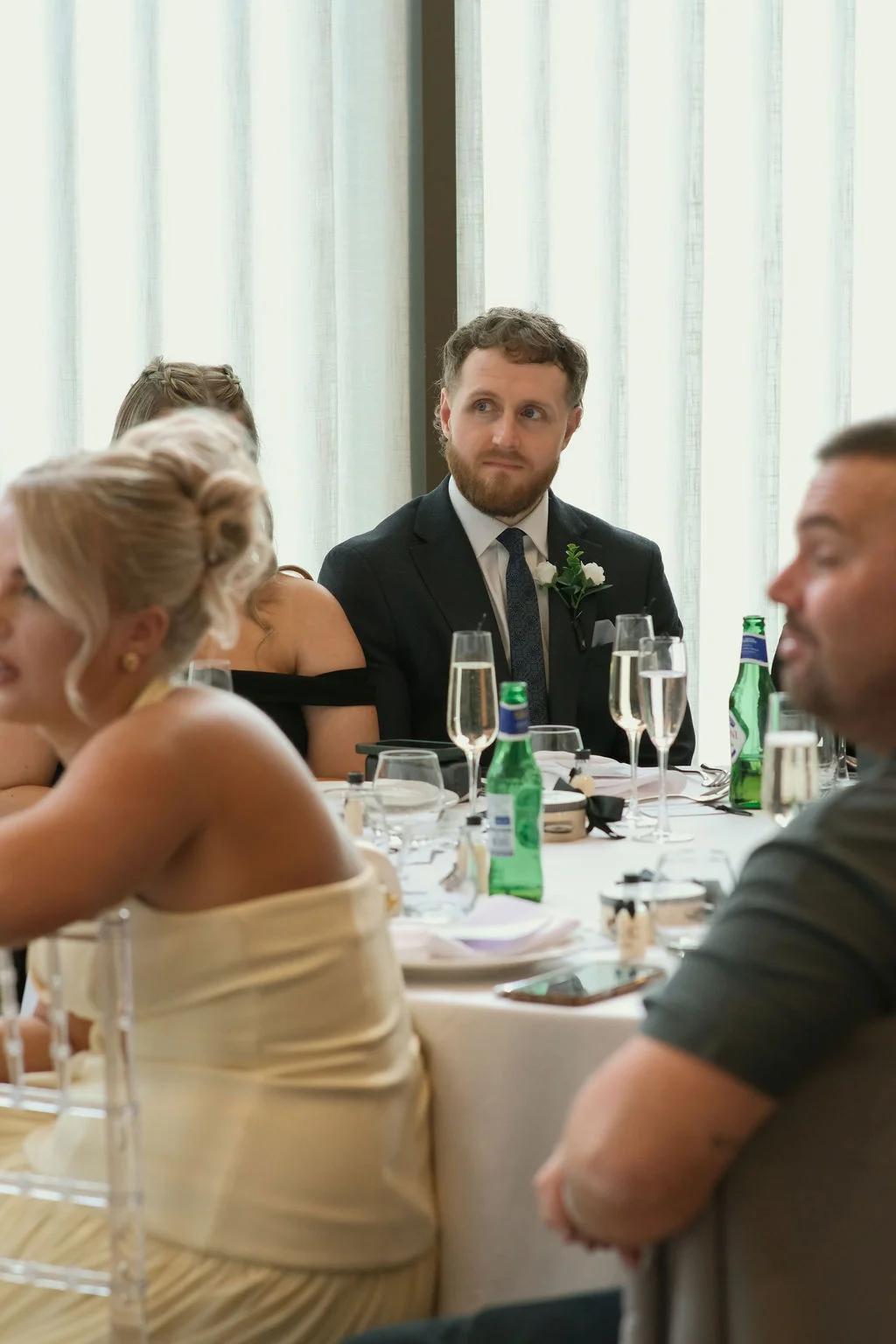 A bearded guest in a dark suit and tie watches attentively during the wedding breakfast while seated at a table with drinks and place settings.