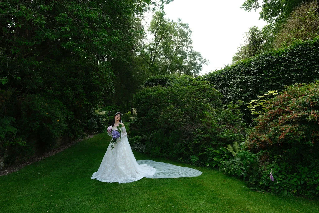 A bride in a lace gown with a long veil stands holding a bouquet on a lush green lawn surrounded by mature garden foliage.