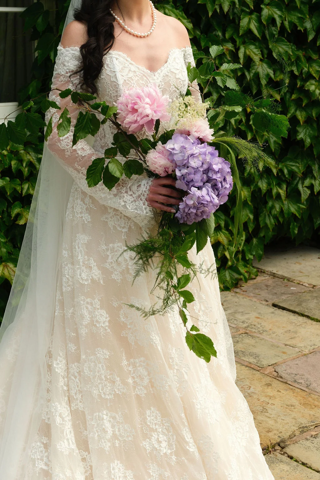 A bride in an off-the-shoulder lace gown with long sleeves holds a cascading bouquet of pink peonies and purple hydrangeas against an ivy-covered wall.
