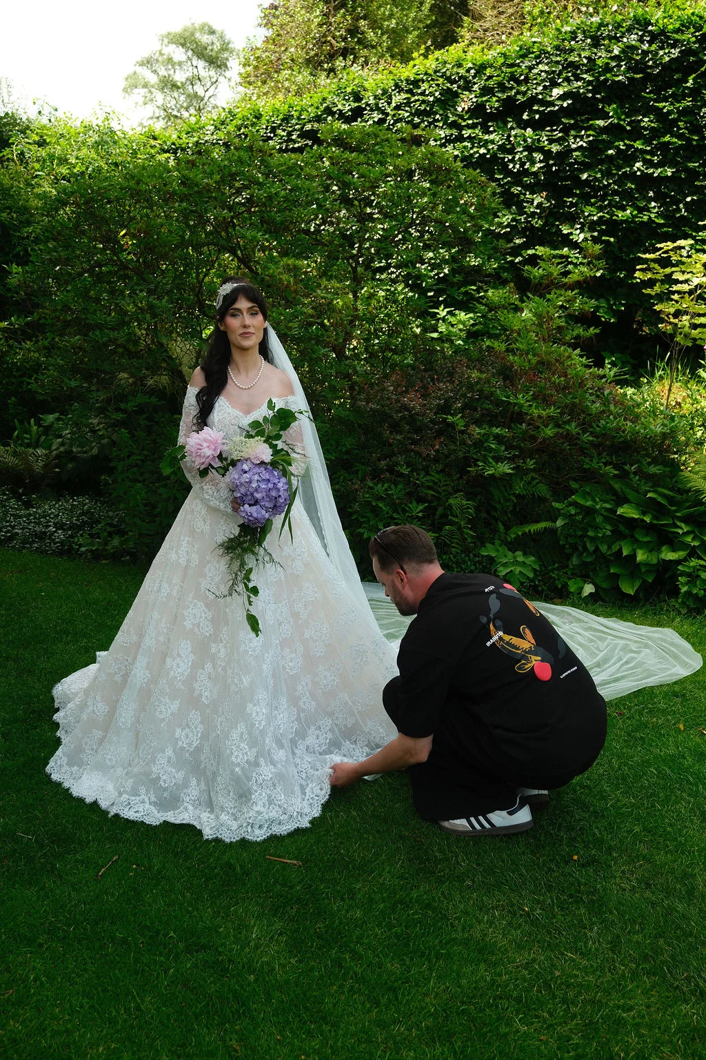 A bride in an off-the-shoulder lace a-line wedding dress with a cascading bouquet stands in a lush garden while someone adjusts her gown.