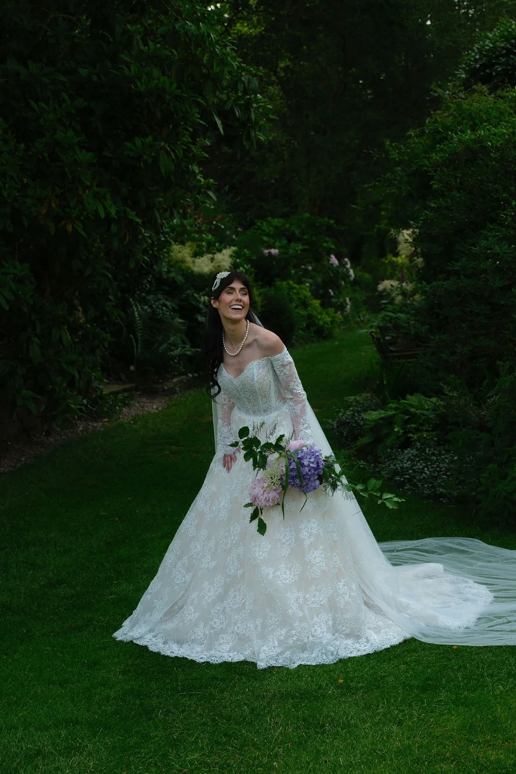 A joyful bride in a lace off-the-shoulder gown laughs while holding a colourful bouquet in a lush green garden.