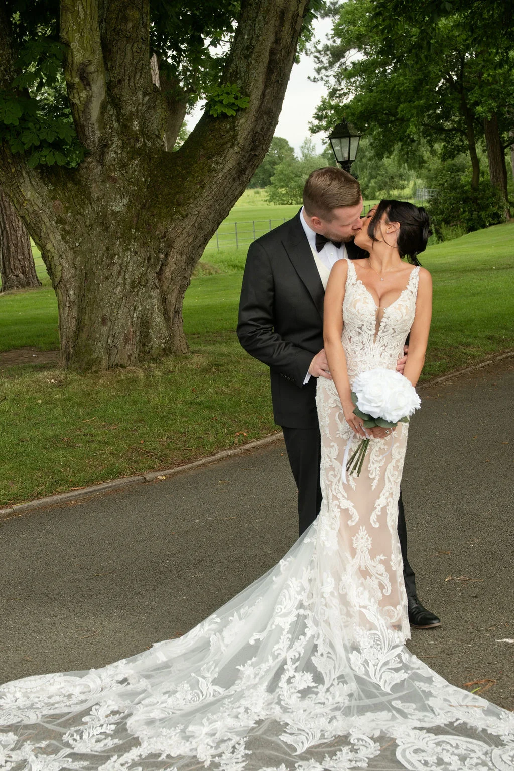 A newlywed couple shares a tender kiss outdoors on a paved pathway beside a large ancient oak tree and manicured lawns.