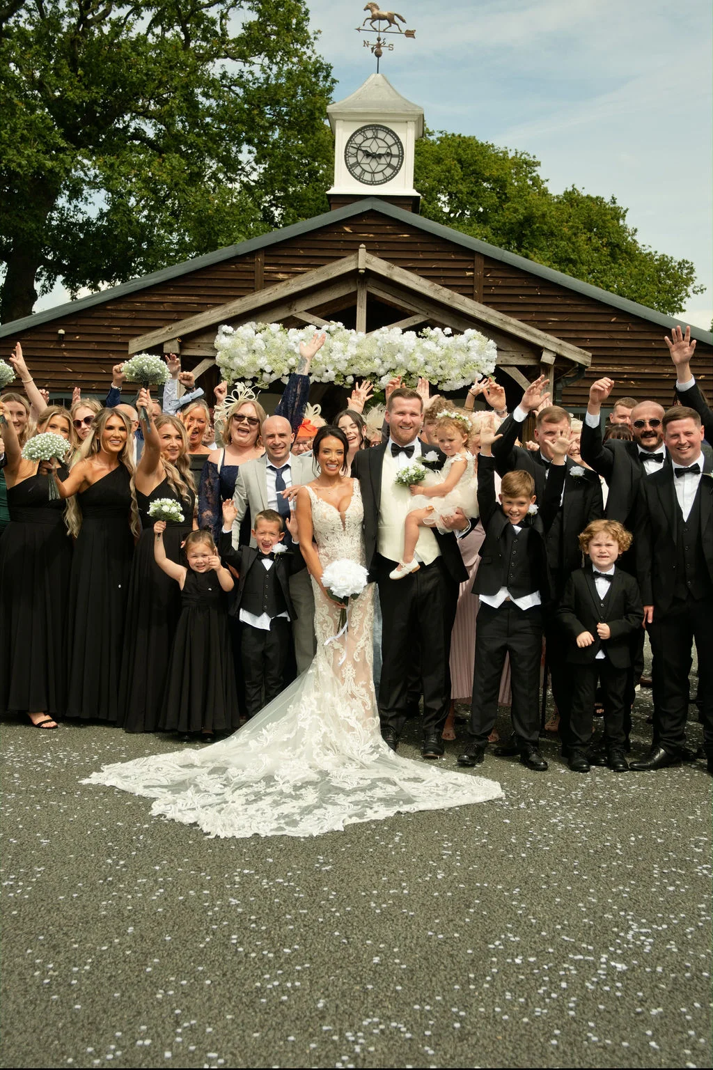 A large wedding party and guests cheer and raise their arms outside a rustic wooden barn venue after a ceremony, with the bride in a lace gown and train holding a white bouquet.