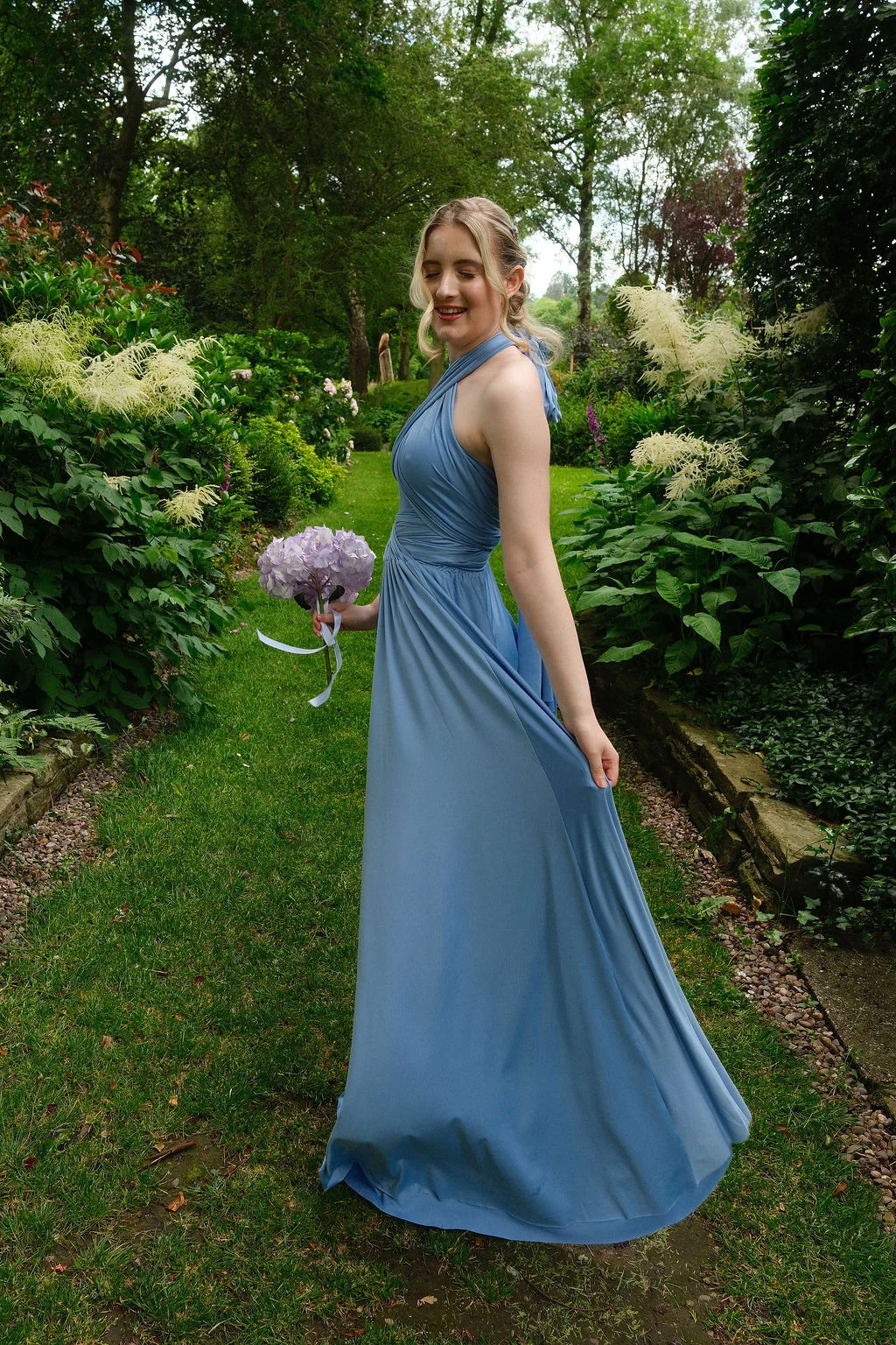 A smiling bridesmaid in a dusty blue halterneck dress poses gracefully along a lush garden pathway while holding a small bouquet of purple flowers.