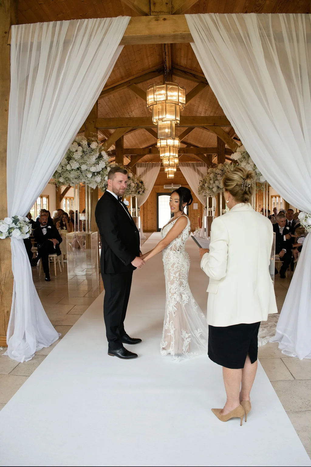 A bride and groom hold hands during their civil wedding ceremony inside a bright converted barn framed by draped white fabric and elegant chandeliers.