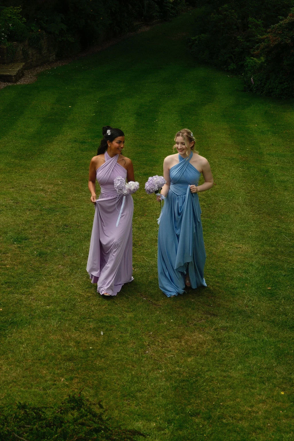 Two smiling bridesmaids wearing halter-neck dresses in lavender and dusty blue walk across a lush green lawn, holding small bouquets.