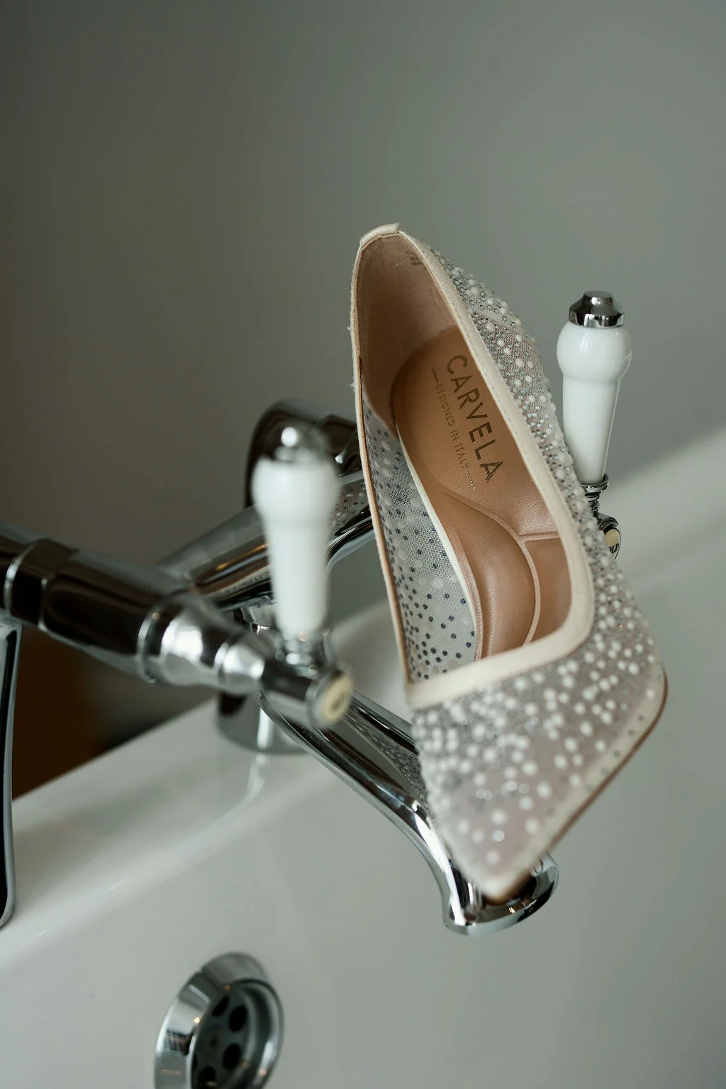 A sparkling pearl-embellished bridal pump rests delicately on the chrome and white ceramic taps of a bathtub.