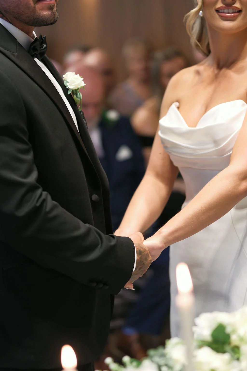 A bride in a draped strapless gown and a groom in a black tuxedo hold hands lovingly during their indoor wedding ceremony.