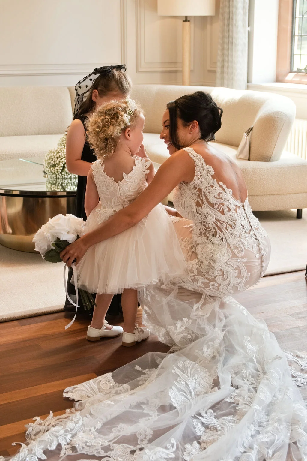 A smiling bride in a detailed lace gown kneels down on wooden flooring to speak warmly with a young flower girl holding a white bouquet.