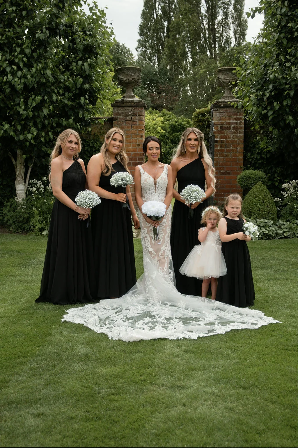 A stunning bride in a plunging lace gown with an elaborate train stands outdoors on a manicured lawn, flanked by bridesmaids in matching black dresses and two flower girls.