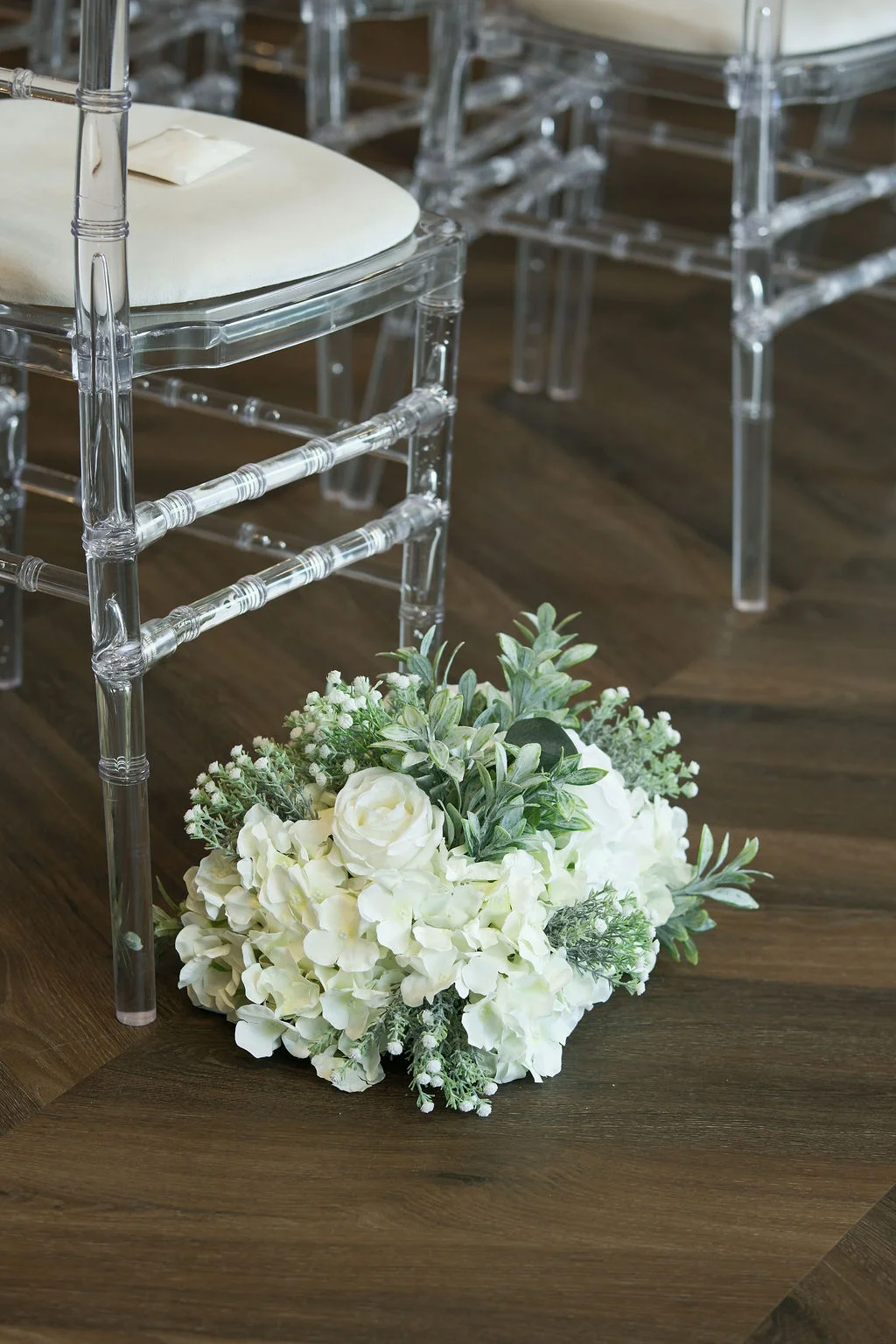 A lush white and green floral arrangement sits on a dark wooden floor beside a clear ghost chair at a wedding ceremony.