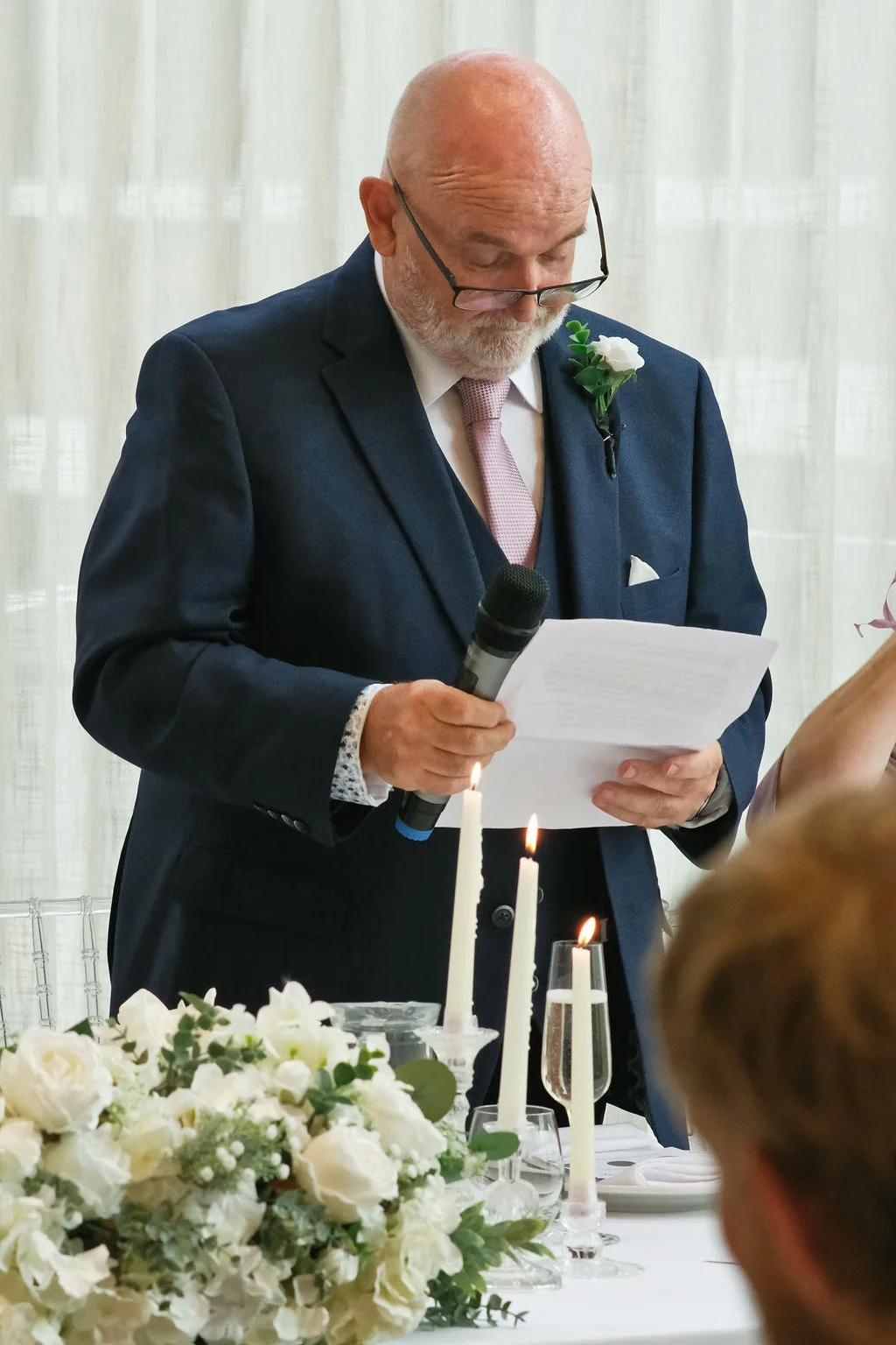 A father of the bride wearing a navy three-piece suit reads a speech from a paper while holding a microphone at the wedding reception table.