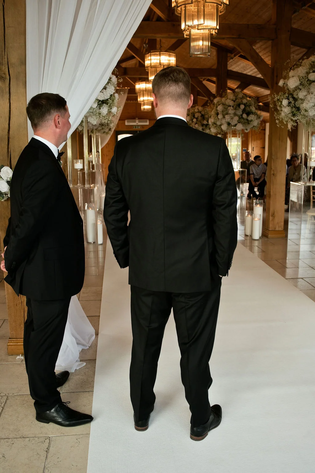 Two grooms in matching black suits stand at the front of a rustic wooden barn venue with a white aisle runner and flickering candles, waiting for the ceremony to begin.
