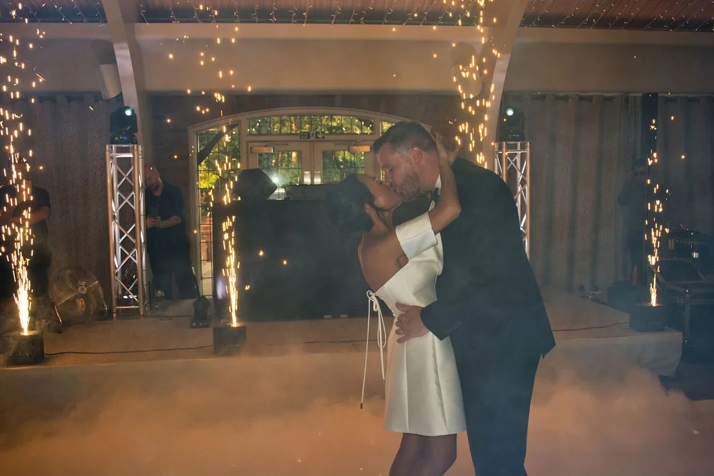 A newlywed couple shares an intimate kiss during their first dance on a dance floor surrounded by dramatic cold spark fountains and low-lying fog.