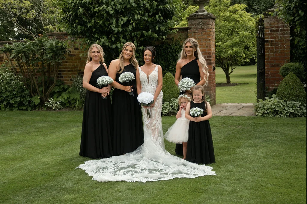 The radiant bride poses gracefully on a manicured lawn with her bridesmaids and flower girls, all wearing striking black dresses against a scenic brick garden backdrop.