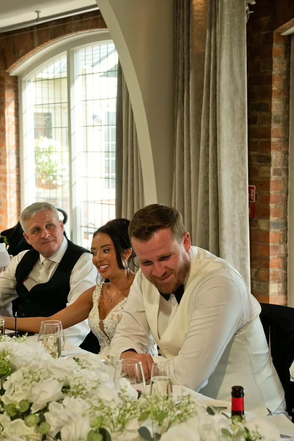 The bride and groom laugh together at the top table during the wedding speeches, surrounded by white floral arrangements and exposed brick.