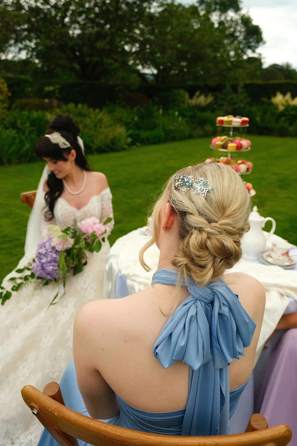 An over the shoulder shot of a blonde bridesmaid in a dusty blue halter dress looking towards a smiling bride holding a bouquet in a lush green garden setting.