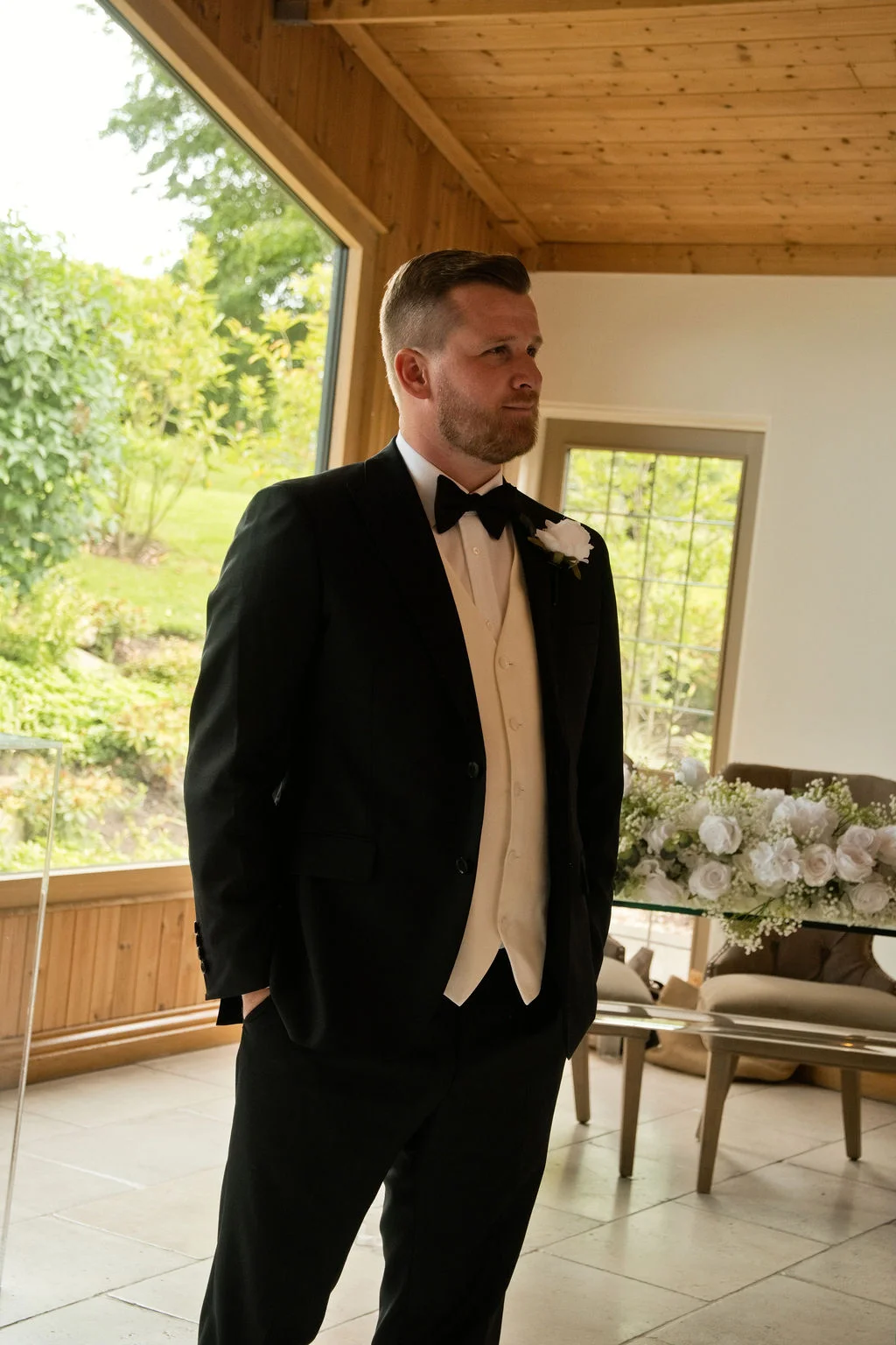 A handsome groom in a black tuxedo and white waistcoat waits patiently inside a sunlit venue with large windows.