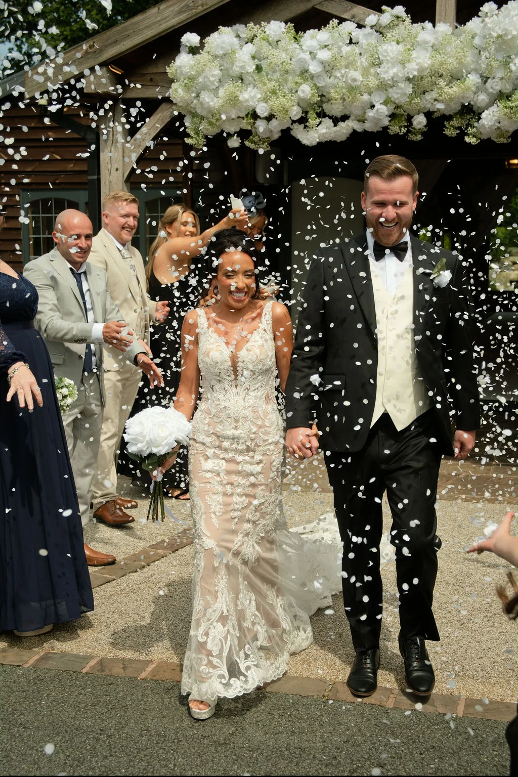 A smiling newlywed couple walks hand in hand through a cascade of white confetti outside a rustic stone barn adorned with lush white floral arrangements.