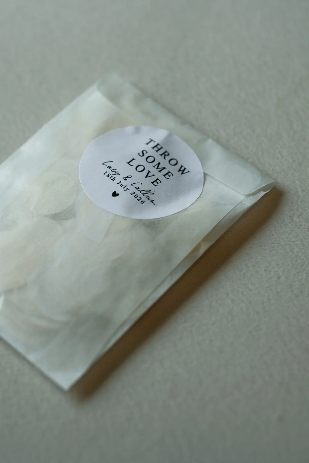 A close-up detail shot of a translucent glassine confetti envelope featuring a round sticker that reads throw some love alongside personalised names and wedding date.