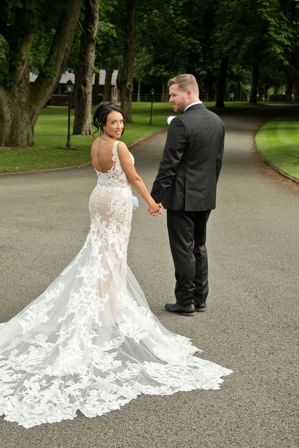 A newlywed couple strolls down a tree-lined driveway, with the bride looking back over her shoulder in a stunning lace gown featuring an open back and a dramatic chapel train.
