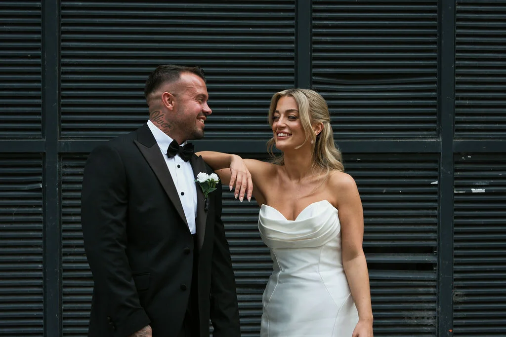 A smiling bride leaning against her groom in a classic black tuxedo in front of a dark slatted background.