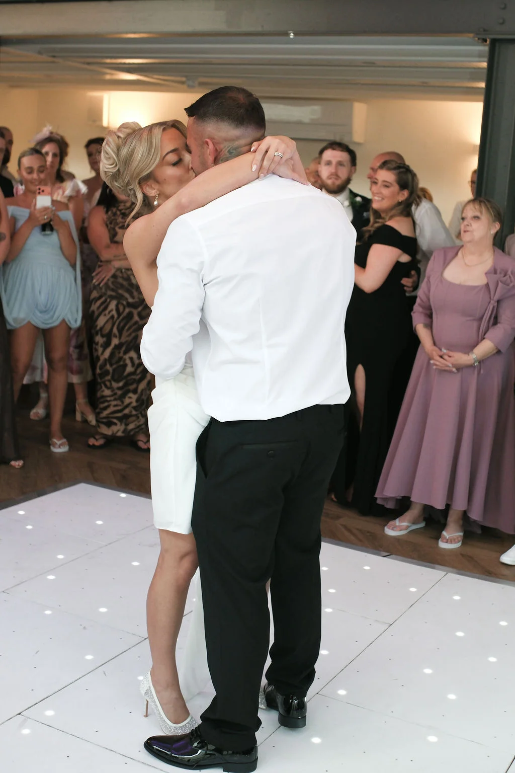 The bride and groom share a passionate kiss during their first dance on an illuminated dance floor while wedding guests watch closely.