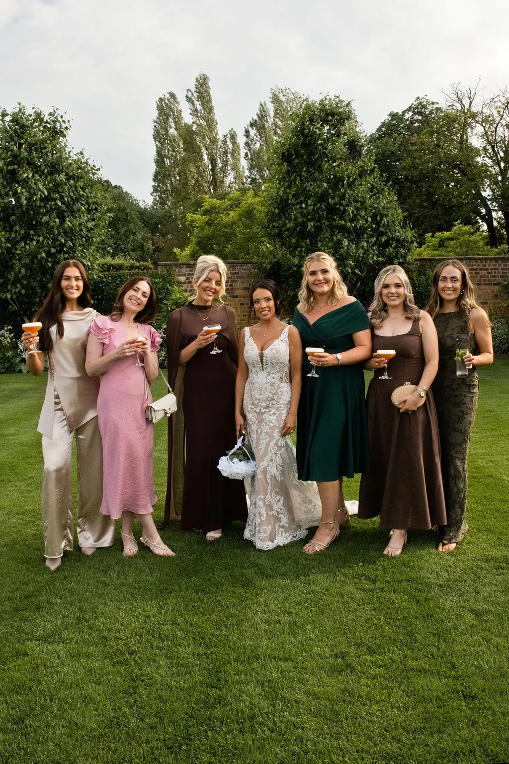 A smiling bride in an intricate lace gown stands proudly on a green lawn with her bridesmaids, who wear mismatched stylish dresses in earthy and pastel tones while holding cocktails.