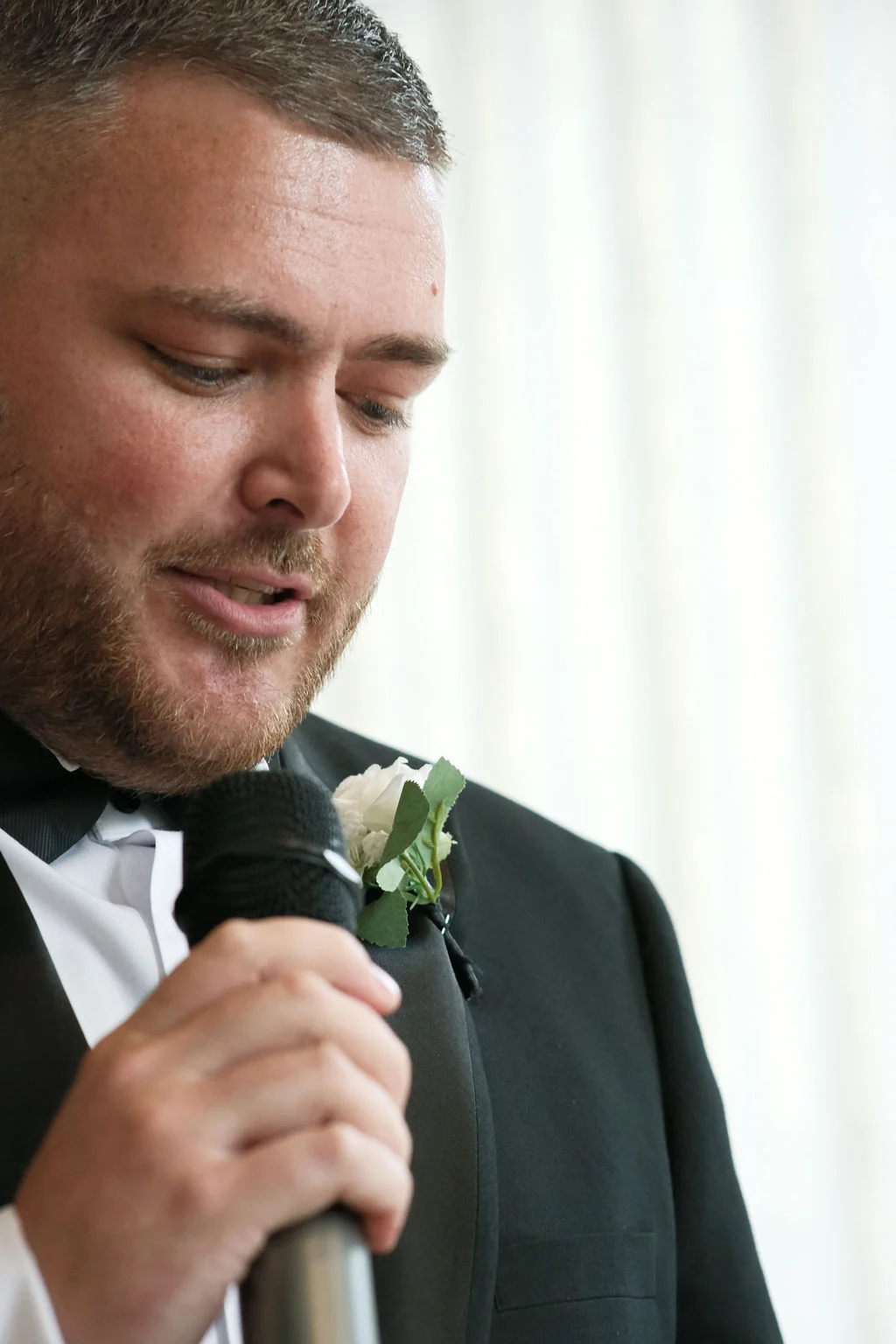 A groom in a classic black tuxedo delivers an emotional wedding speech while holding a microphone and wearing a white rose boutonniere.