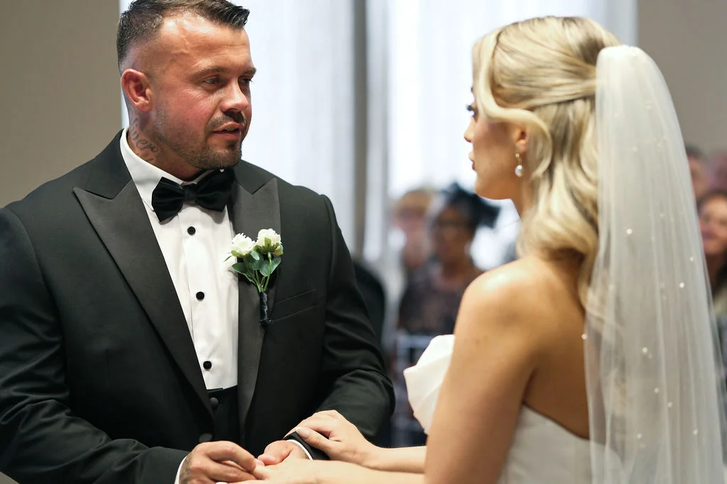 A groom in a black tuxedo and bow tie places a ring on the bride's finger while exchanging vows during their wedding ceremony.