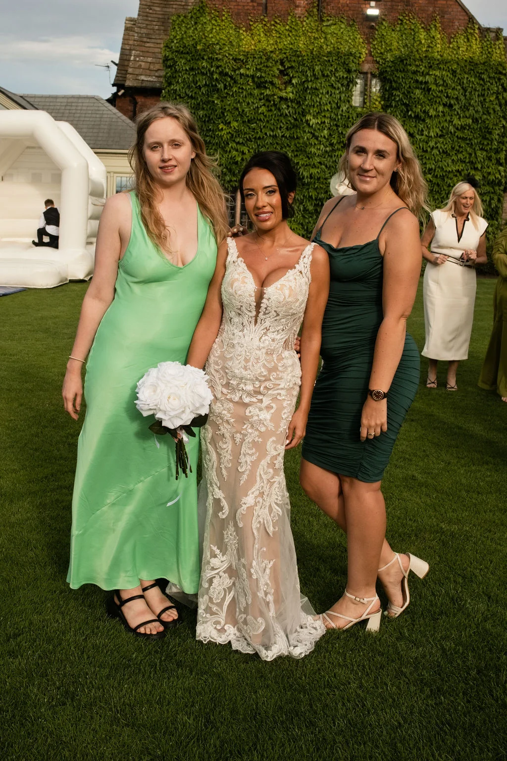 A smiling bride in an intricate lace gown stands outdoors on a lawn, flanked by two bridesmaids wearing vibrant green dresses.