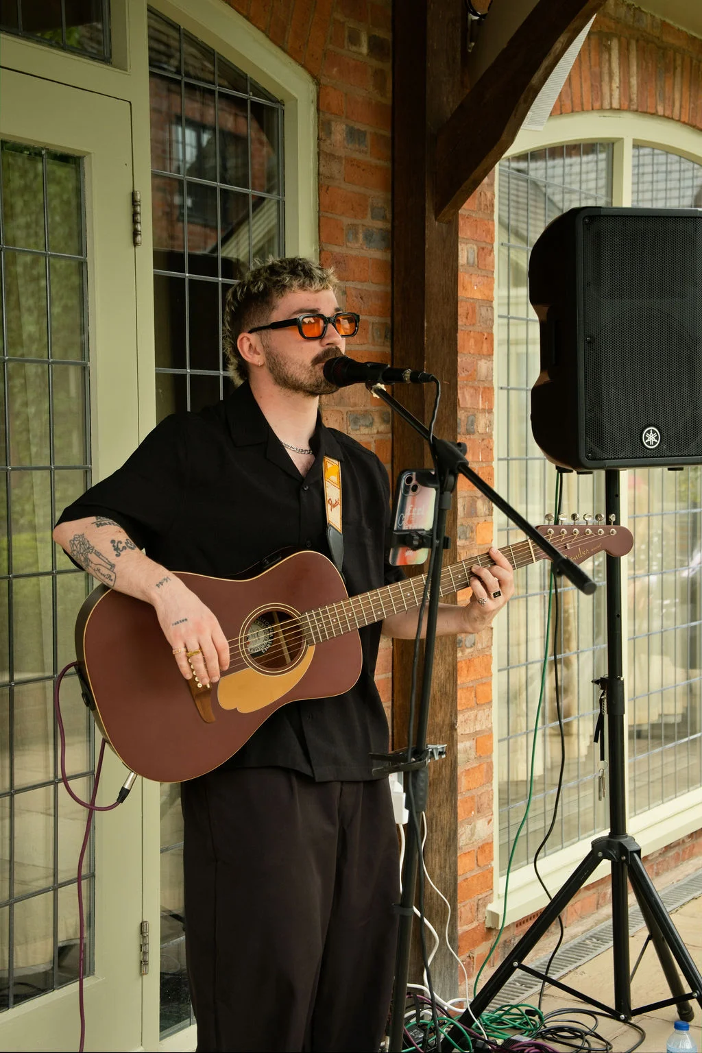 A stylish male singer with tattoos and tinted sunglasses performs live with an acoustic guitar in front of a brick wall and large windows.