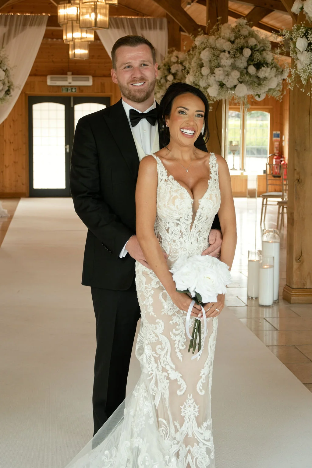 A smiling bride holding a white rose bouquet stands with her groom inside an oak-framed barn decorated with hanging floral arrangements and warm candlelight.