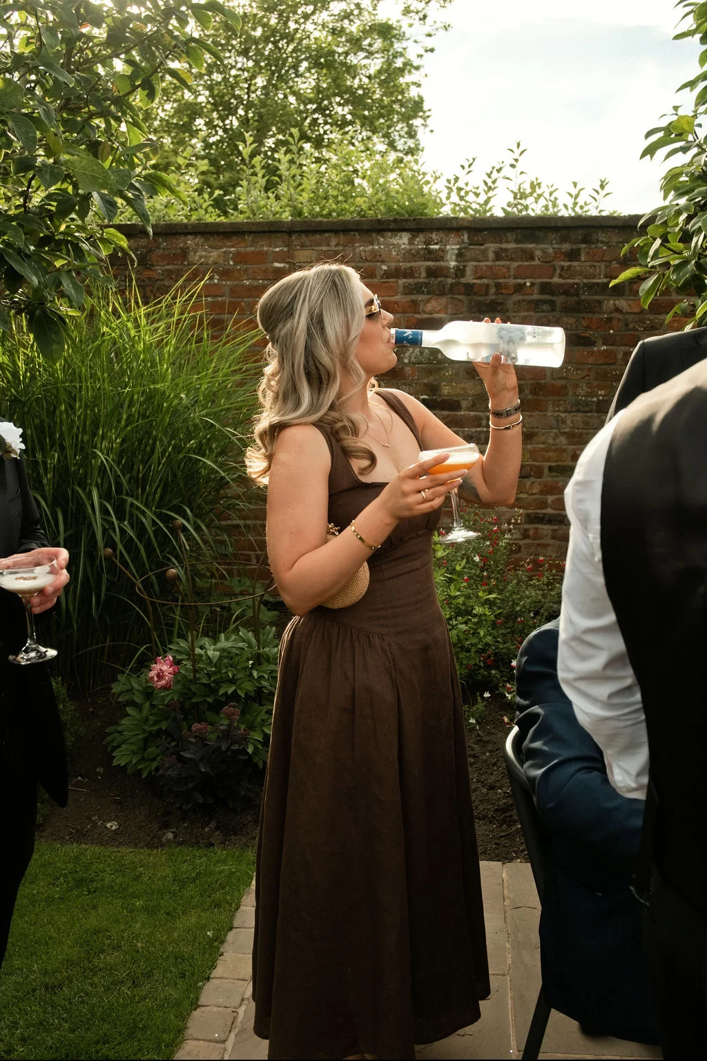 A wedding guest in a brown dress drinking straight from a bottle of vodka during an outdoor garden reception.