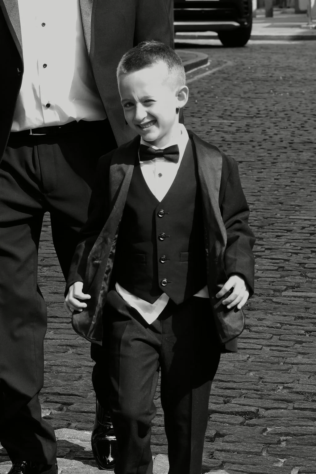 A young page boy wearing a sharp tuxedo and bow tie laughs joyfully while walking across a cobbled courtyard alongside an adult.