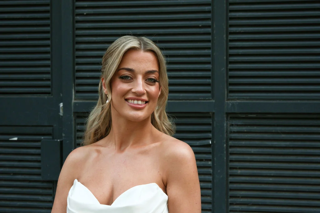 A smiling bride wearing a strapless gown with a sweetheart neckline and a half-up hairstyle poses against a dark slatted background.