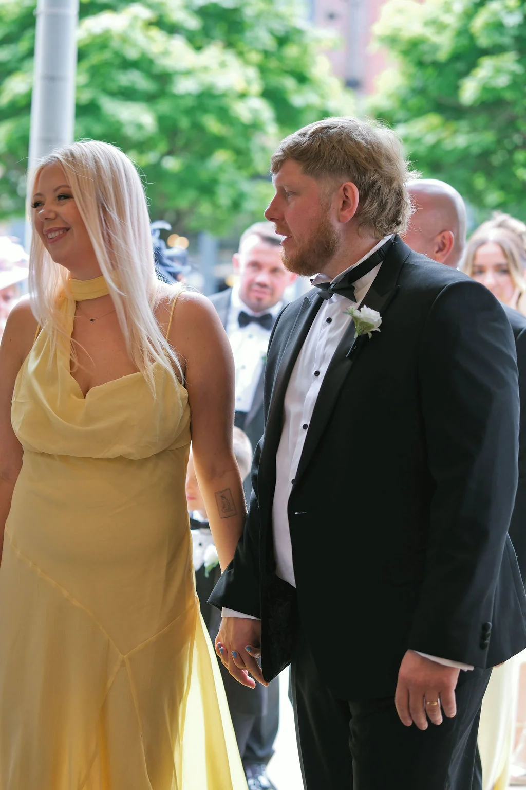 A smiling blonde bride in a yellow dress holding hands with the groom in a classic black tuxedo during a wedding celebration.