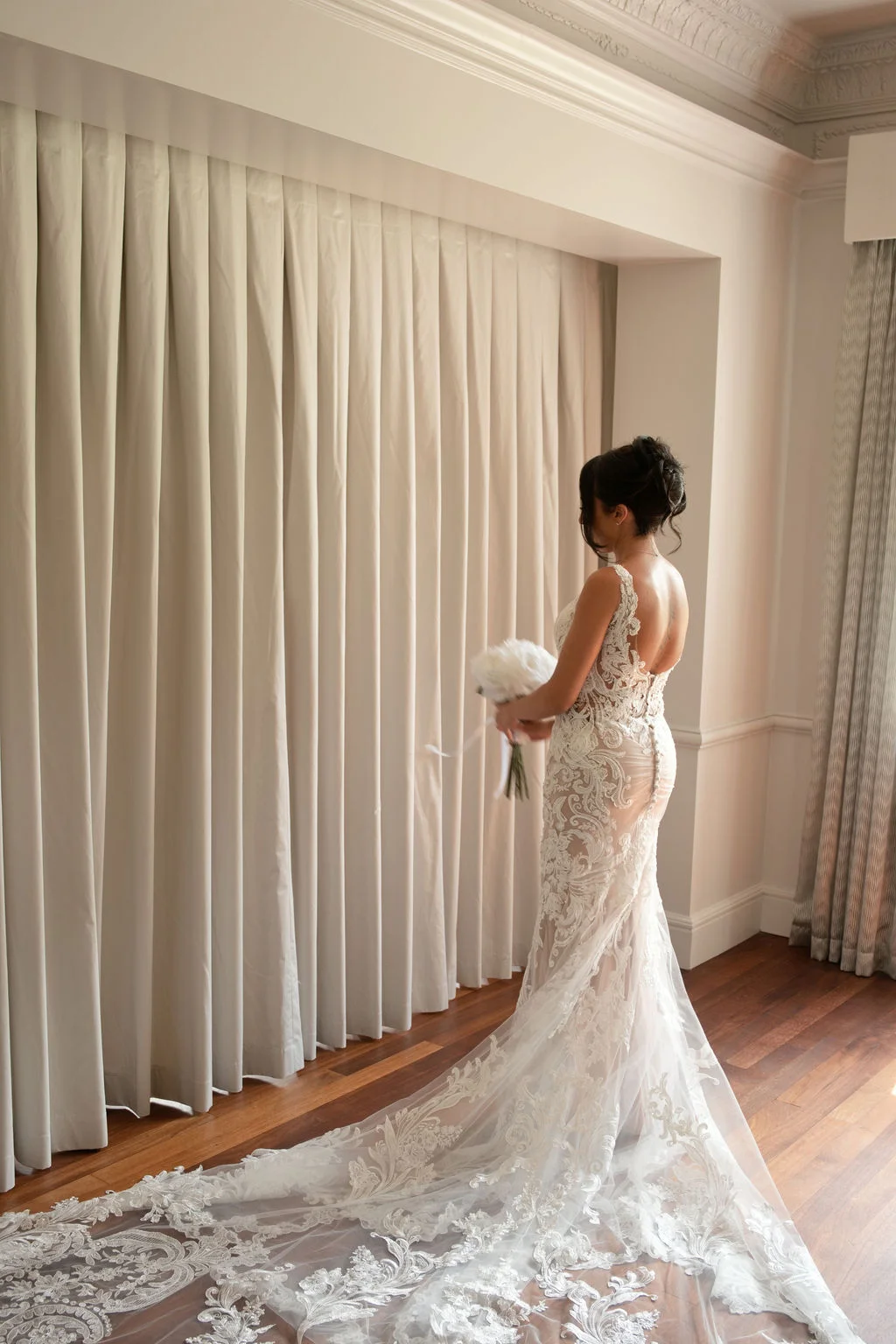 A bride stands by a draped window holding a white bouquet, wearing an intricately patterned lace wedding dress with a long train.