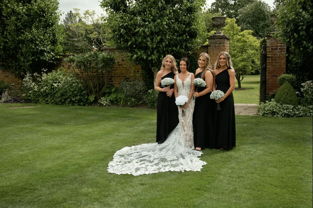 A stunning bride in a plunging lace mermaid gown with a long embroidered train poses on a manicured lawn with her four bridesmaids in matching black one-shoulder dresses.