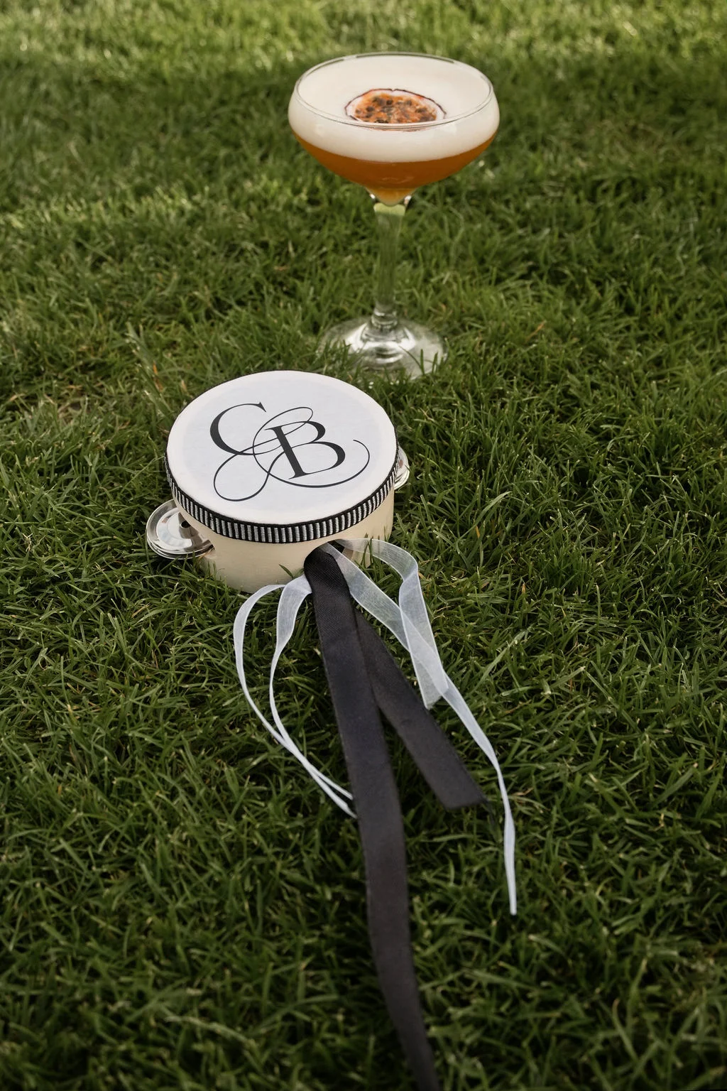 An elegant tambourine featuring a couple's monogram and black and white ribbons rests on lush green grass alongside a passionfruit cocktail in a coupe glass.