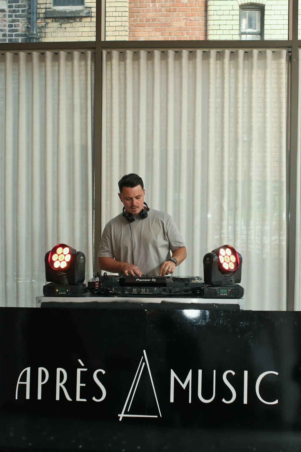 A wedding DJ focused on mixing tracks at a branded booth with professional lighting set up against a backdrop of floor-to-ceiling windows and exposed brick.
