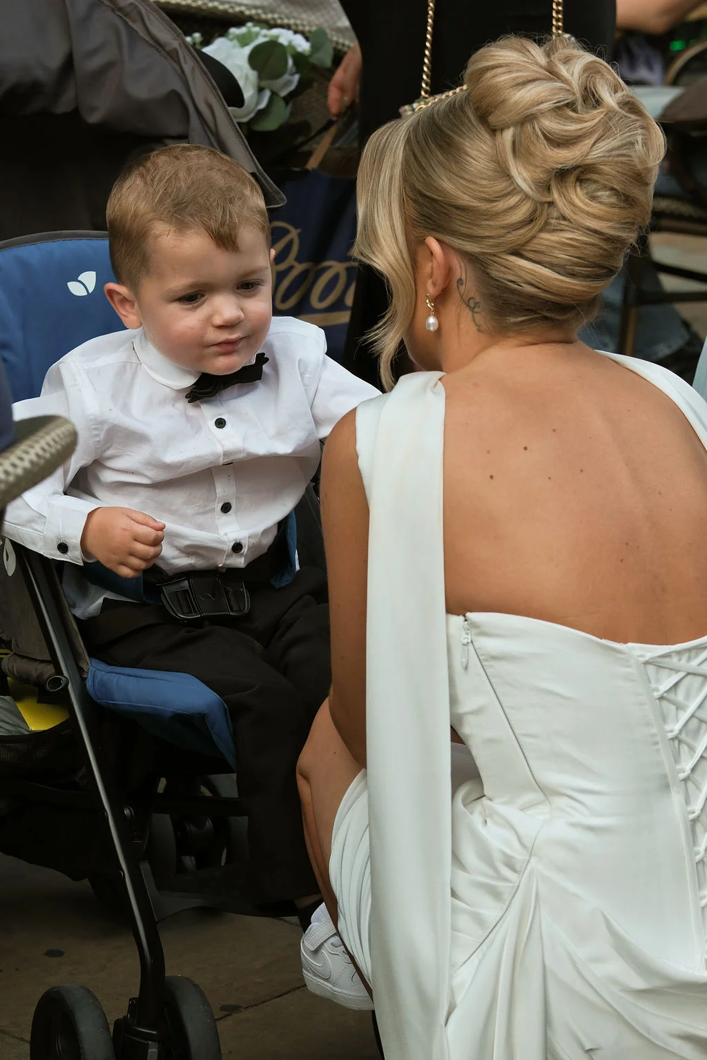 A blonde bride wearing a structured white gown with a lace-up back crouches down to lovingly interact with a young page boy sitting in a pushchair.