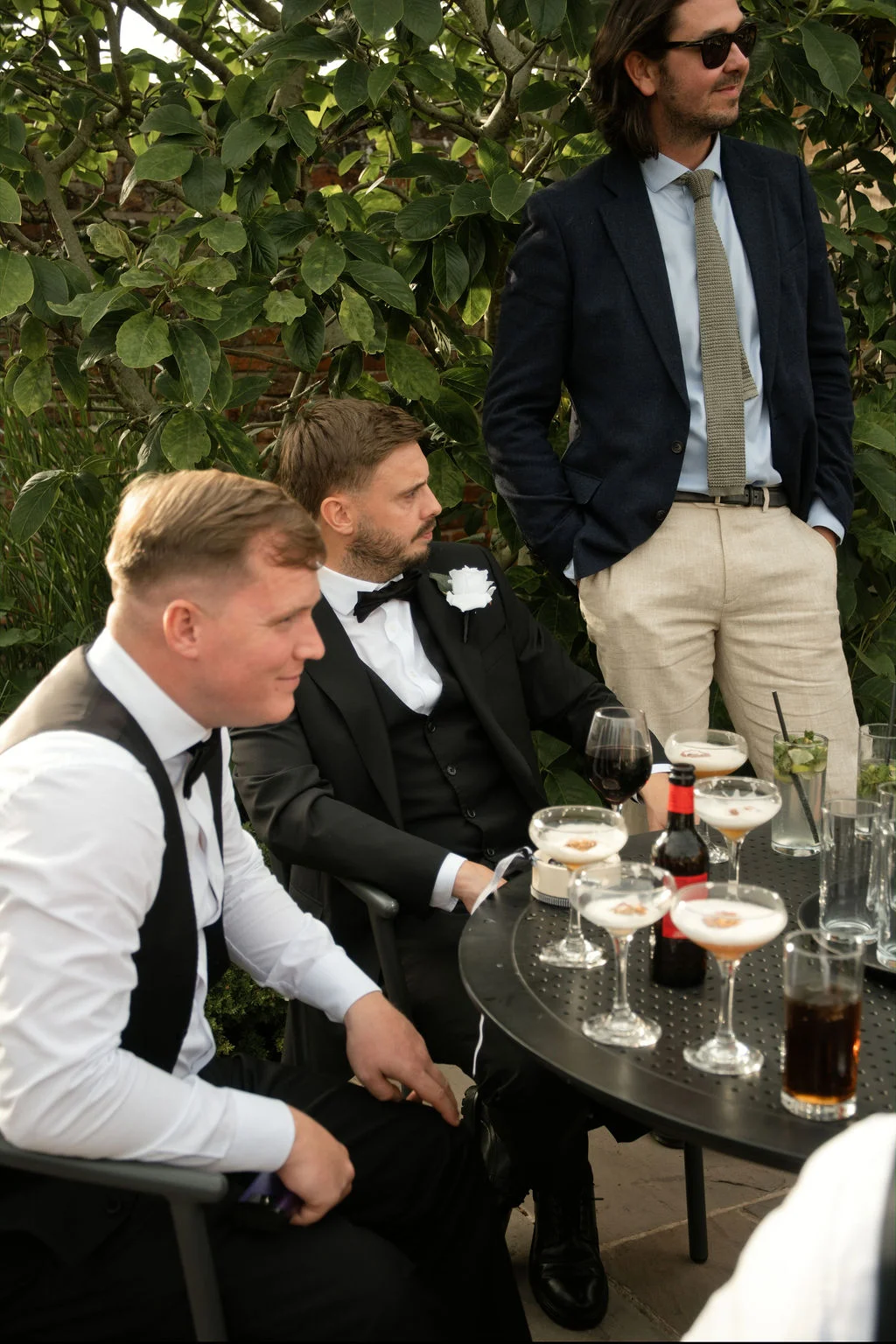 A group of groomsmen and wedding guests relax and socialise outdoors around a table laden with cocktails during the drinks reception.